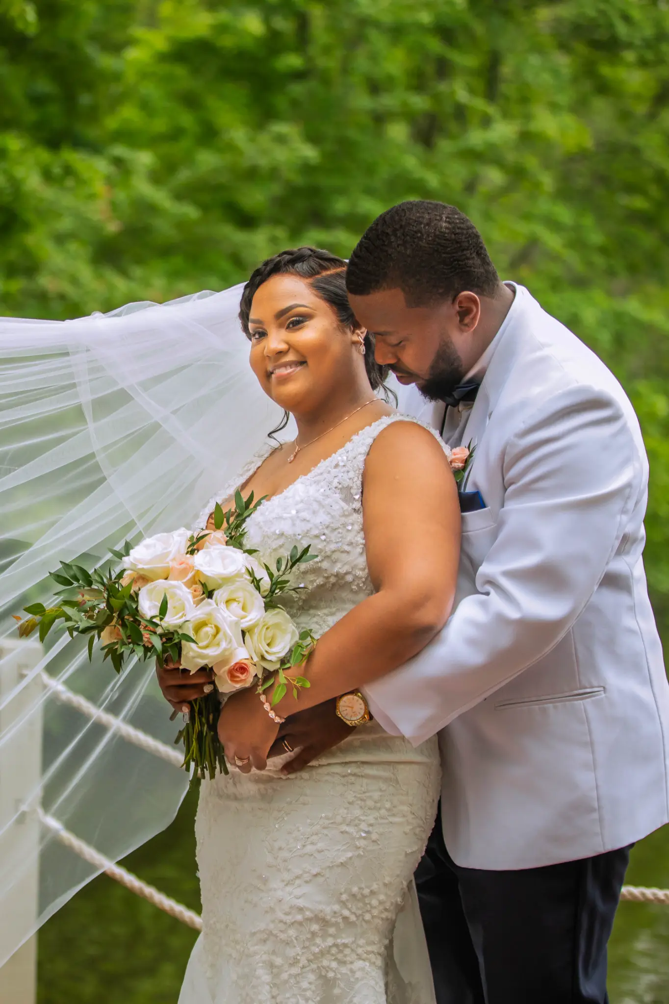 Black couple sharing an intimate outdoor portrait — groom in white tuxedo, bride with flowing veil and peach bouquet